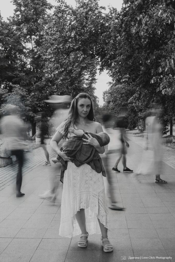 Мајка дои бебе во јавен простор, стоејќи мирно во парк додека луѓе се движат околу неа – симболична црно-бела фотографија што ја истакнува интимноста на доењето во хаотичниот урбан живот.